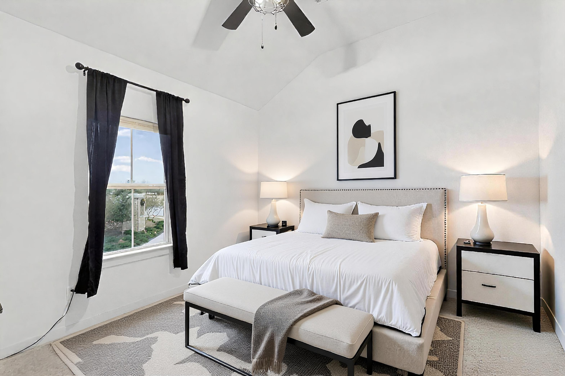 Secondary bedroom with black curtains, framed art above the bed, white bedding and lamps on both sides.