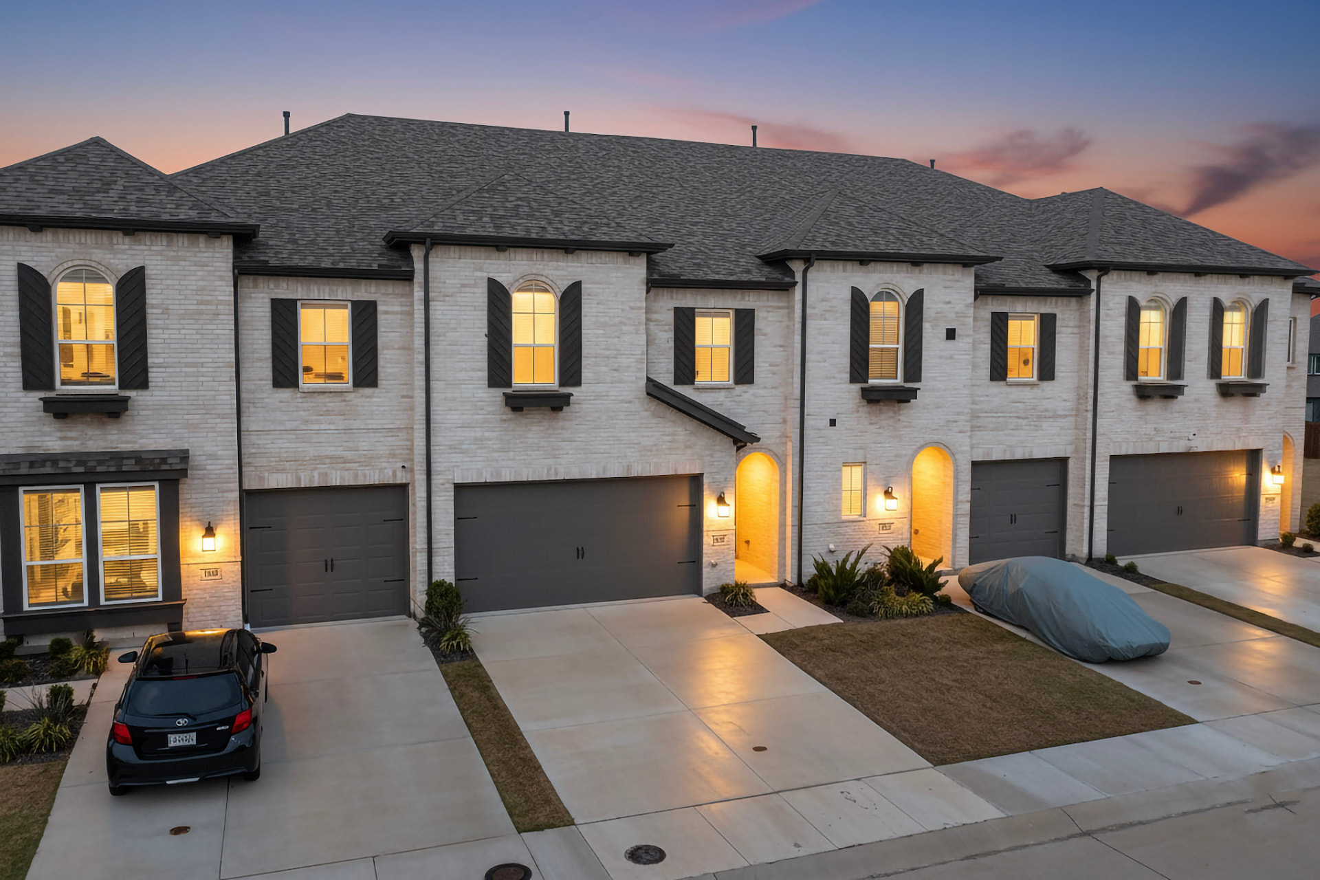 Brick townhome row at twilight with warm lit windows, entry lights and a sunset sky.
