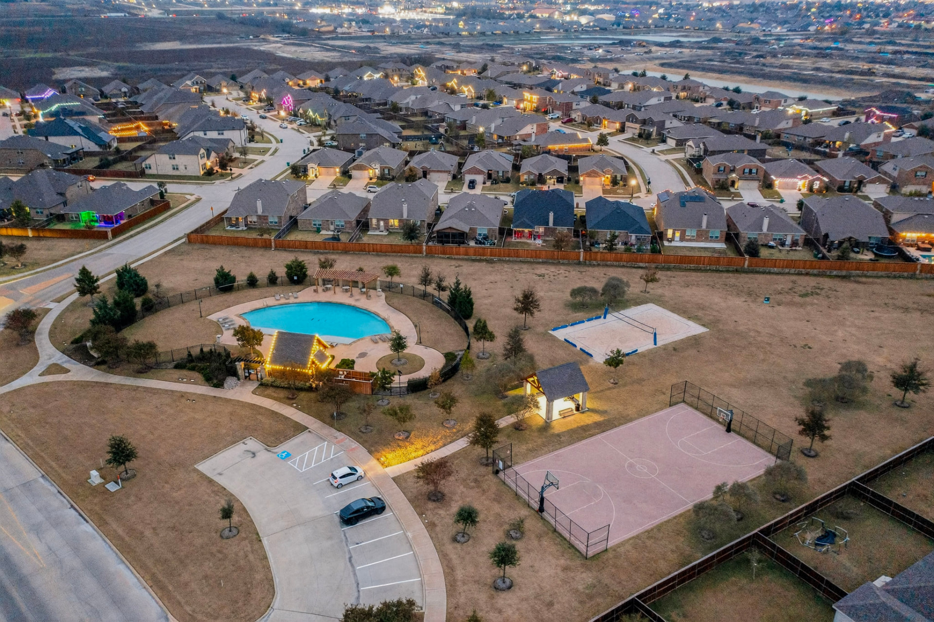 Aerial view of the neighborhood at dusk with the community pool lit, volleyball and basketball courts and pavilion.